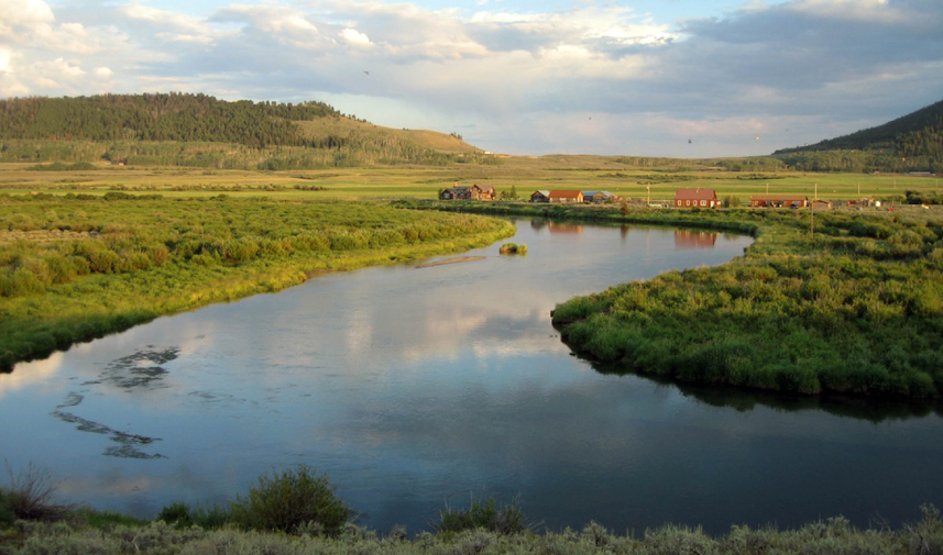 A river winds its way through a ranch with small houses dotting the coastline and rolling hills in the background