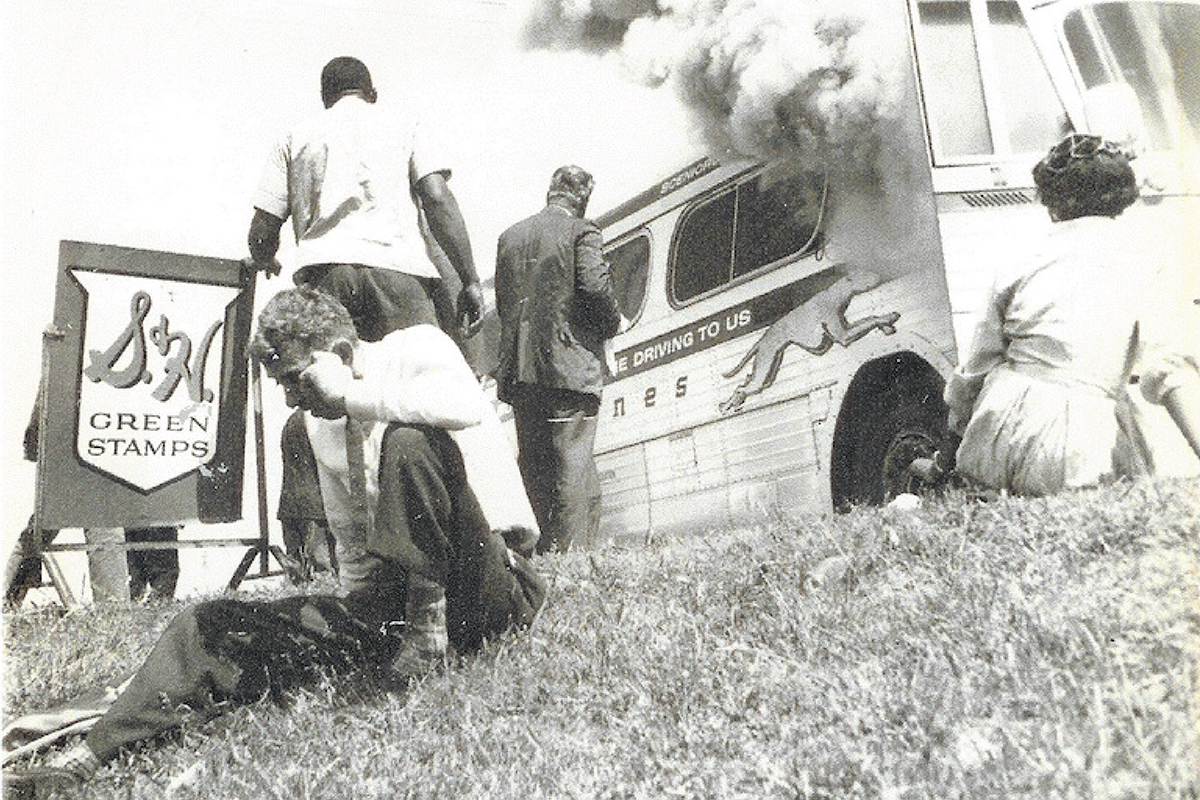 Old black and white photograph of people standing and sitting outside of a smoking bus