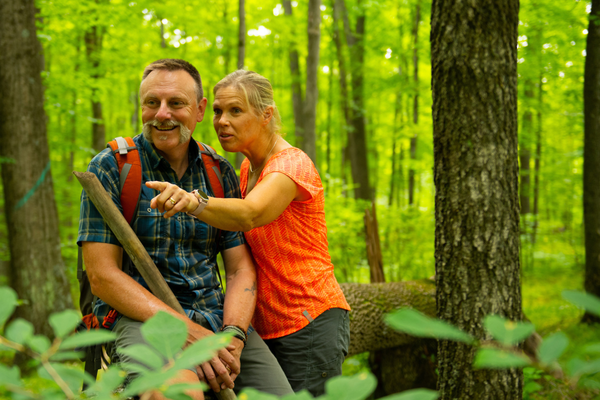 A woman pointing to something in the forest, next to a man smiling and holding a walking stick