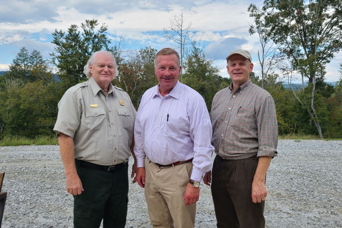 Three men standing on a gravel walkway, smiling at the camera, with trees in the background