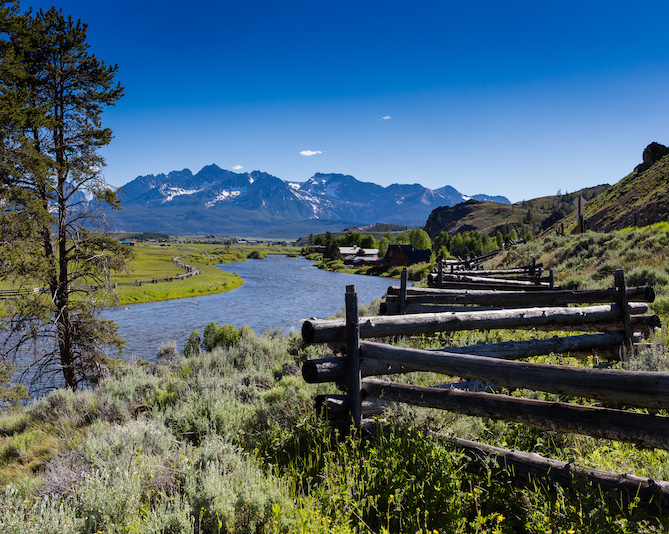 Log fence in a grassy hillside next to a river with mountains in the background