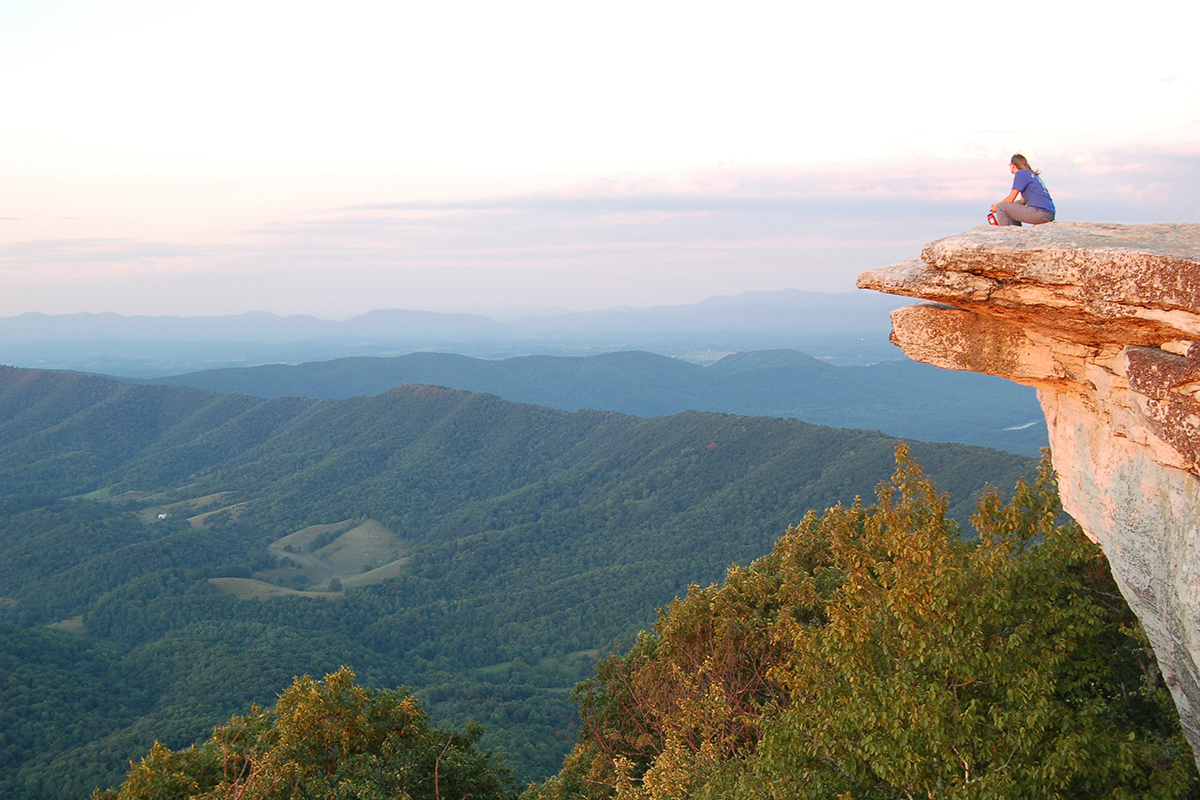 Woman sitting on a cliff edge overlooking a green valley at sunrise