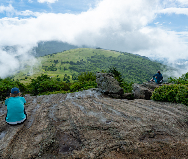 Two people sitting on a cliffside looking over a green valley